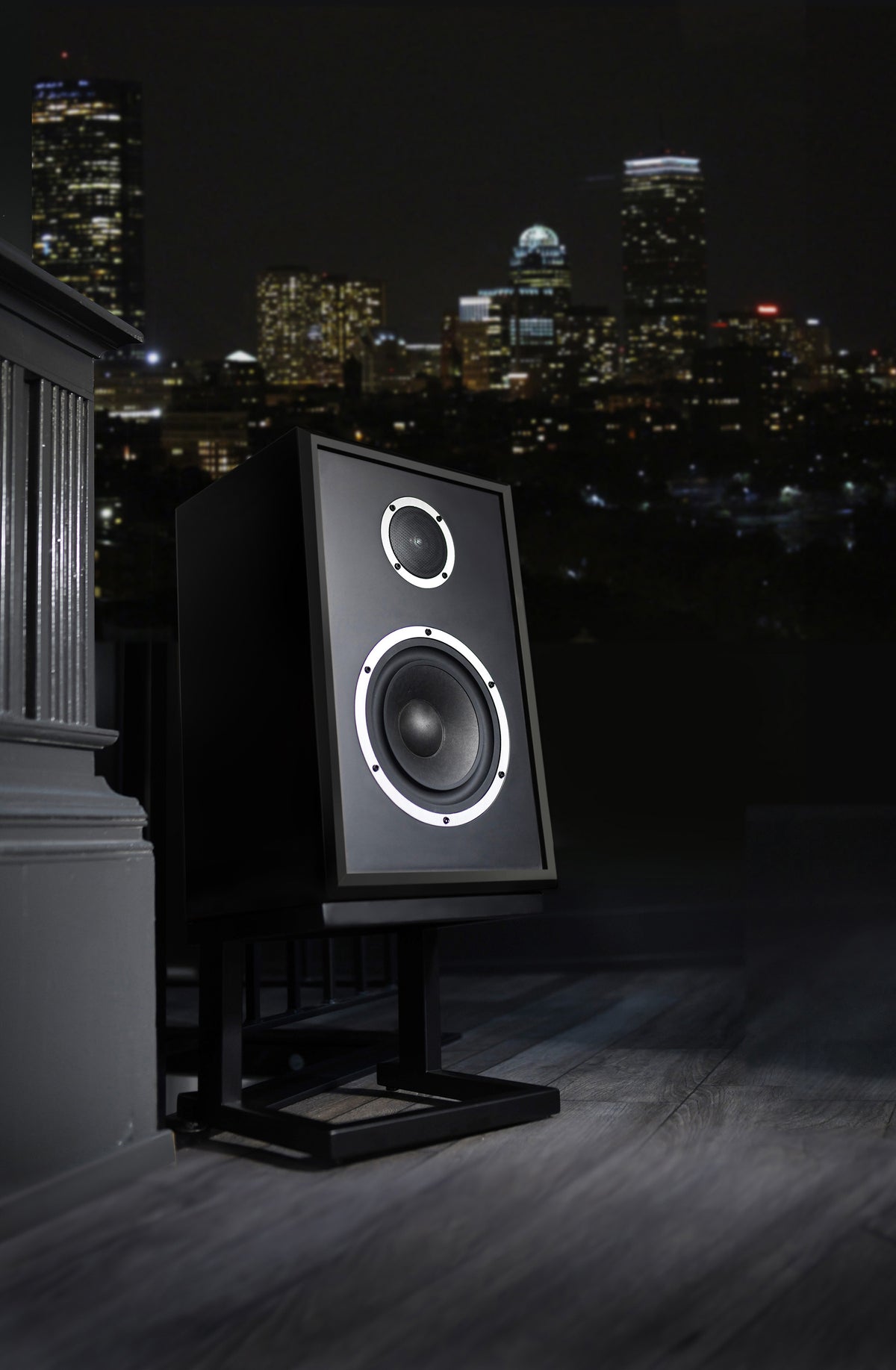 A black studio monitor speaker on a stand, photographed outdoors at night with a city skyline in the background.
