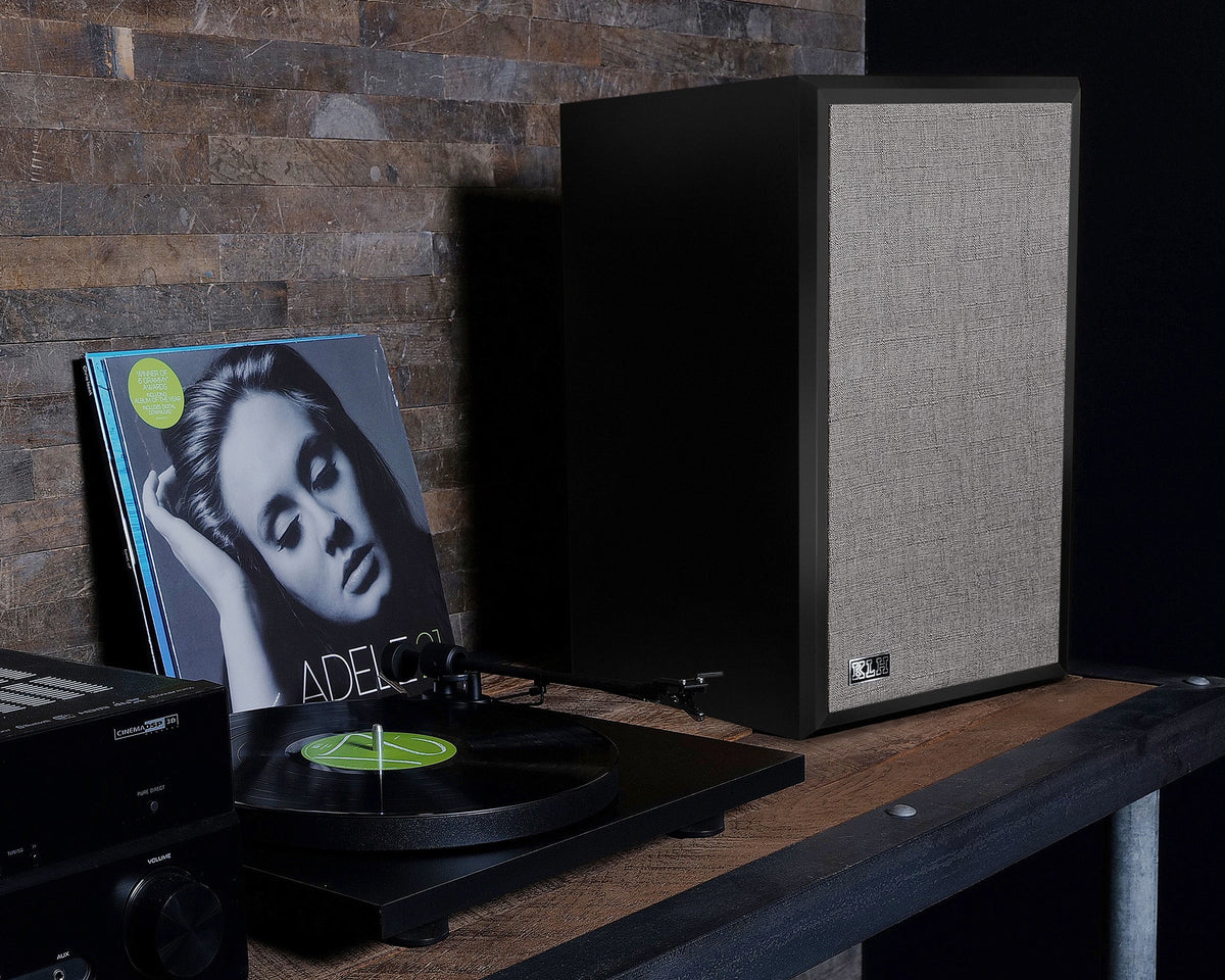 A black speaker with a beige woven grille stands on a rustic wooden table next to a turntable, vinyl record, and audio equipment against a brick wall background.