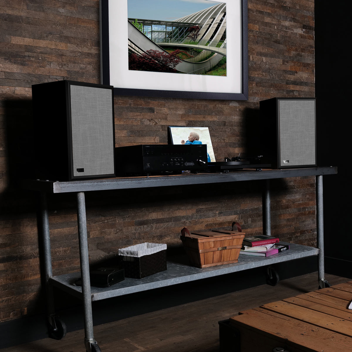 A pair of black speakers with gray fabric grilles sit on a dark metal rolling media console against a rustic wood-paneled wall. A record player, framed art, and storage baskets are arranged on and below the console.