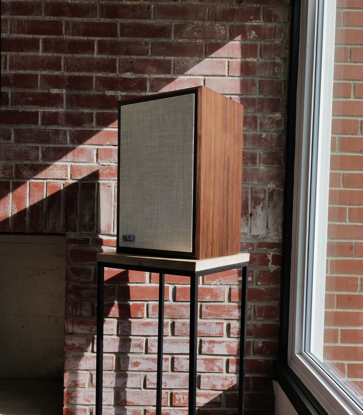 A vintage-style wooden speaker with a beige fabric grille sits on a black metal stand in front of a red brick wall, with strong sunlight casting shadows.