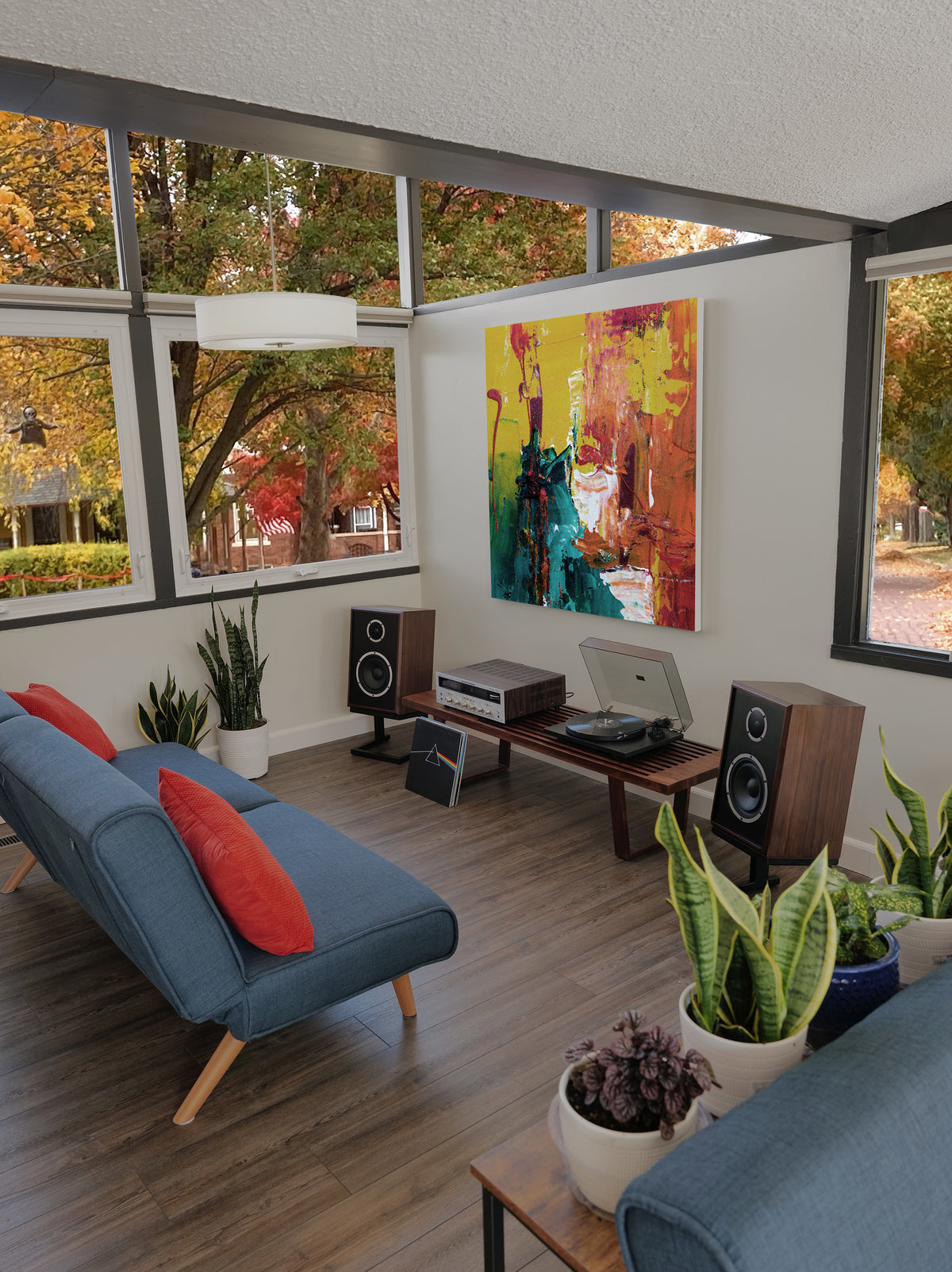 A modern living room with a blue sofa, red pillows, wood flooring, large windows with fall trees outside, and a colorful abstract painting on the wall.