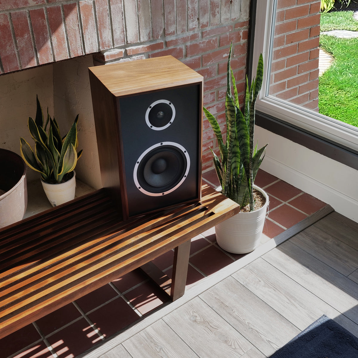 A wooden speaker cabinet with a black front grille sits on a wooden bench in a sunlit room with brick walls, potted plants, and a window view of grass.