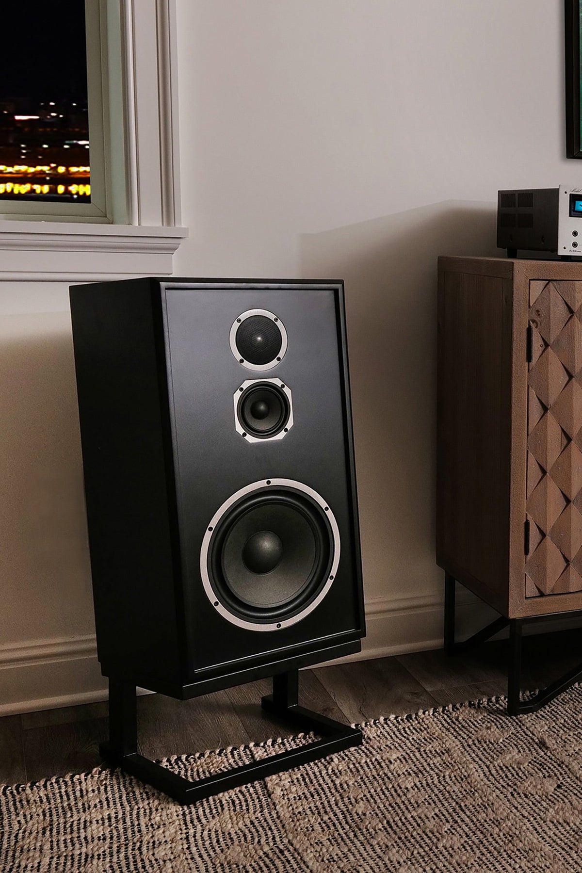A black bookshelf speaker on a metal stand in a home theater setup next to a wooden cabinet with vinyl records.