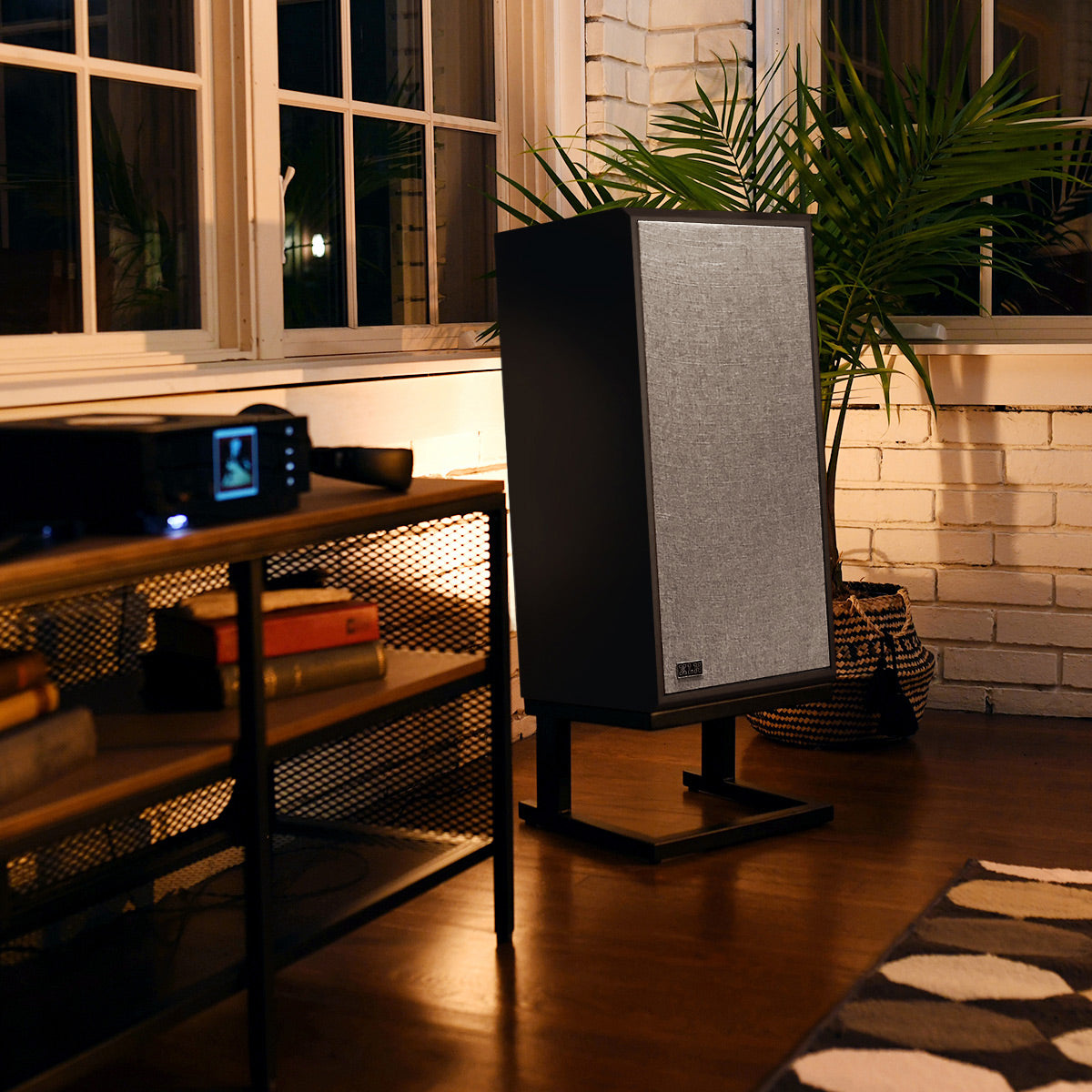 A tall floor-standing bookshelf speaker with a textured gray front grille and black side panels, displayed on a black metal stand in a cozy, warmly lit room with a brick wall, potted plant, and wooden floor.