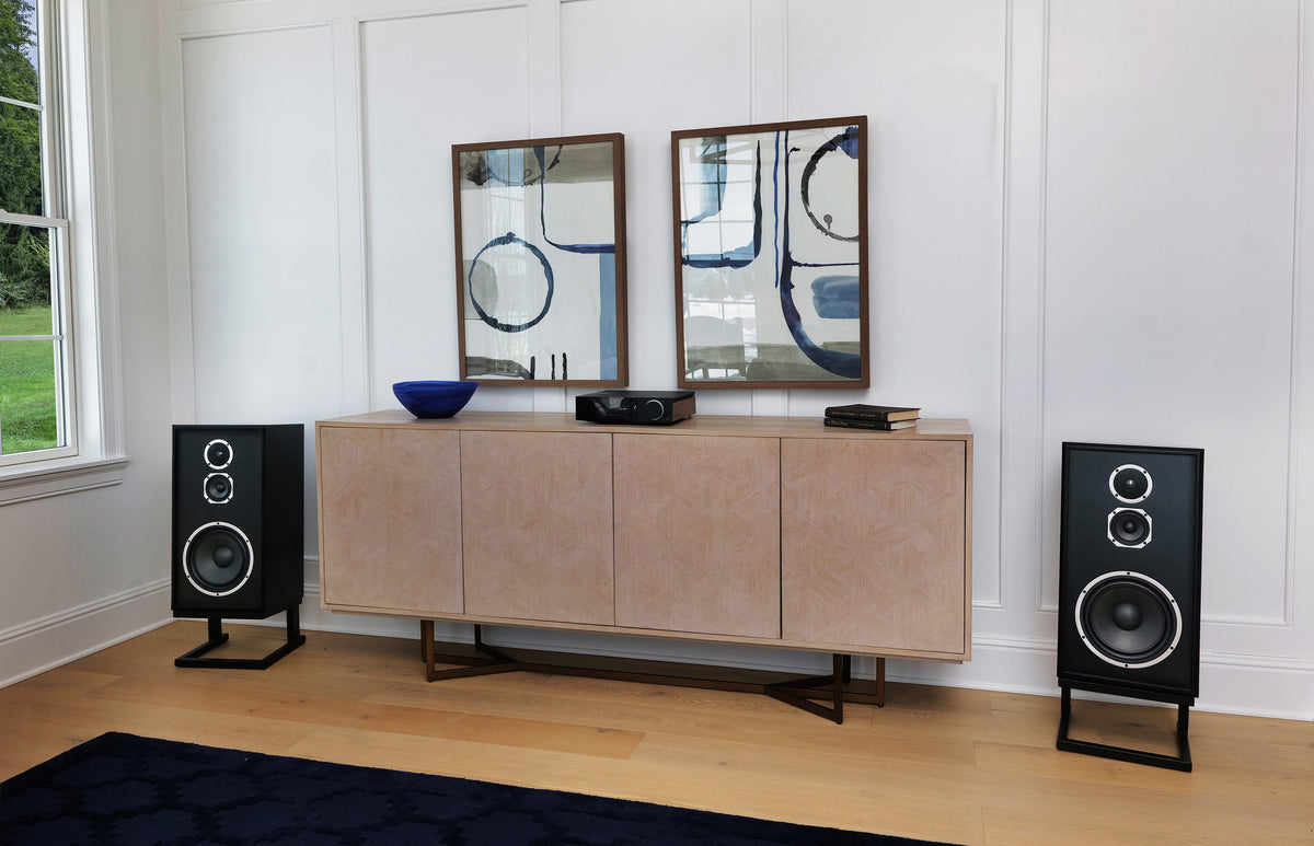 A mid-century modern sideboard cabinet in a light beige textured finish with dark wood legs, styled in a bright room with two black floor speakers on either side, two abstract framed mirrors, and a dark blue bowl on top.