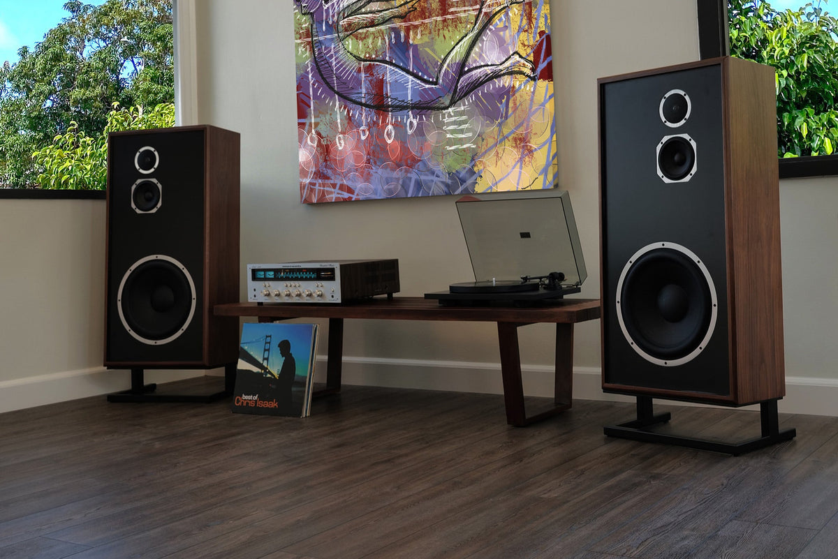 A modern room with wood flooring, large windows, and a colorful abstract wall painting. Two tall wood speaker towers are placed on either side of a small console table holding a vinyl record player and receiver.