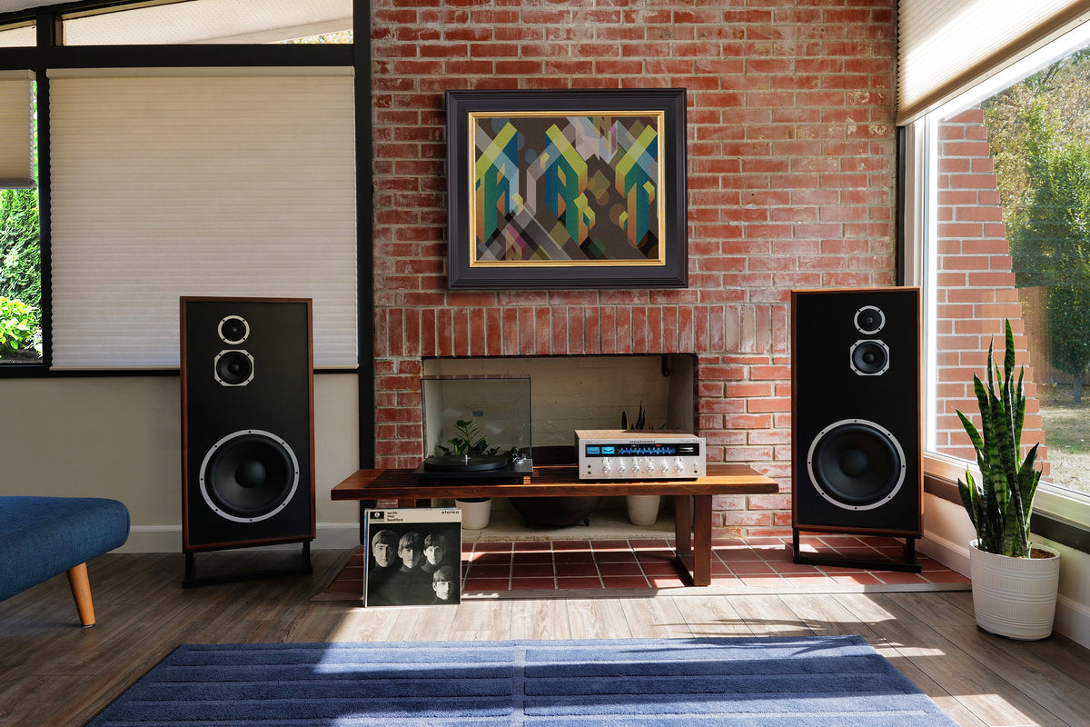 A pair of black tower speakers with wood-grain side panels are displayed in a modern living room setting on either side of a brick fireplace. The room features a blue rug, a wooden console table, and a colorful abstract painting on the brick wall.