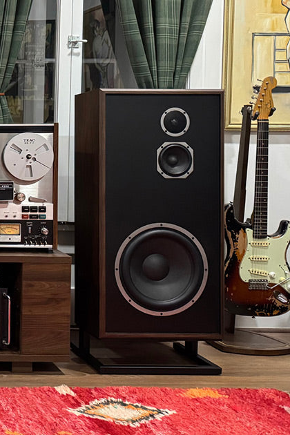 A tall floorstanding bookshelf speaker with a dark wood cabinet, black front baffle, and dual silver-finished tweeters, shown in a styled room with guitars, a reel-to-re recorder, and a red rug.