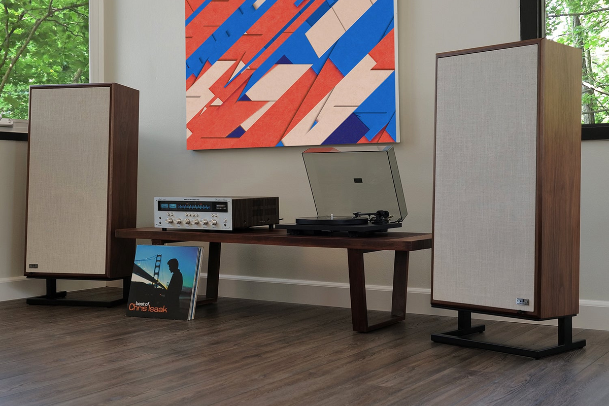 A pair of tall floor-standing speakers with wood-grain fronts and beige fabric cones, flanking a wooden console table in a modern room. A turntable, receiver, and vinyl records sit on the table beneath a colorful abstract painting.