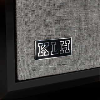 Close-up of a speaker grille with a black rectangular badge featuring the letters K, L, and H in white, mounted on a gray woven fabric cover.