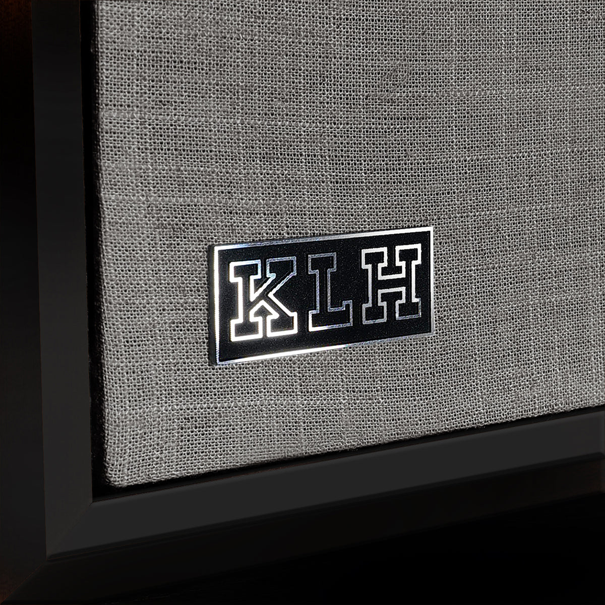 Close-up of a speaker grille with a black rectangular badge featuring the letters K, L, and H in white, mounted on a gray woven fabric cover.