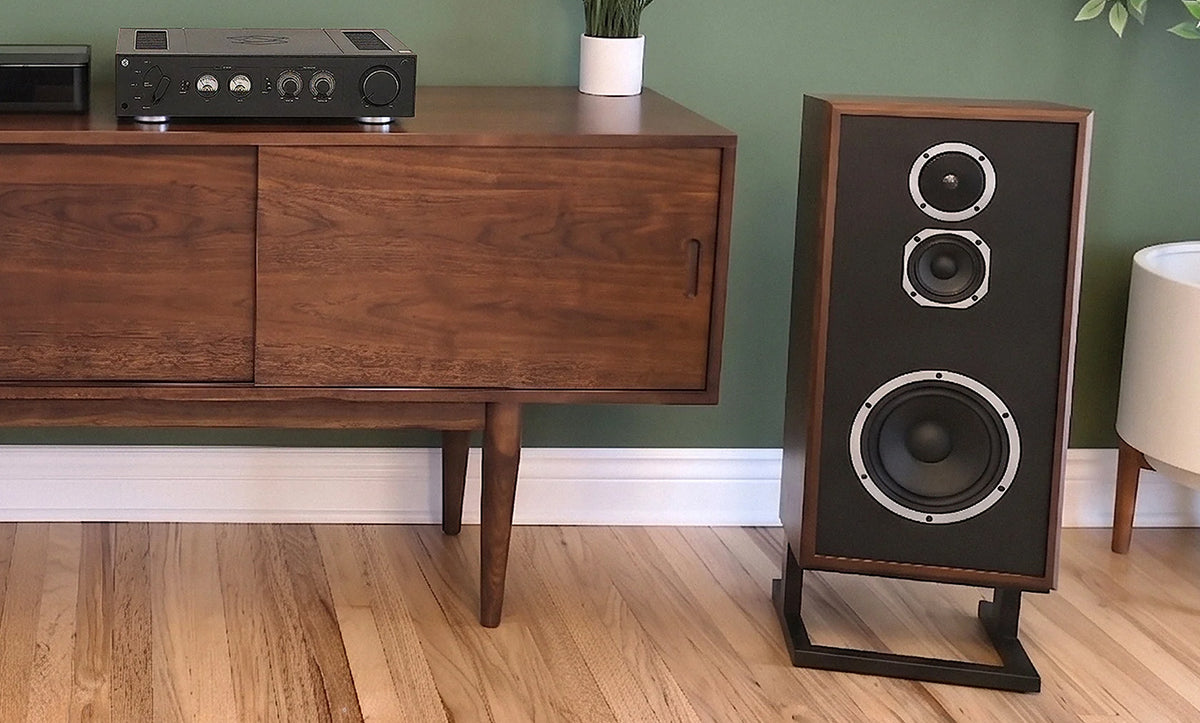 A tall wooden floor-standing speaker with a black front grille and silver speaker cones stands on a black metal stand next to a mid-century modern wooden credenza in a room with a green wall and wood floors.